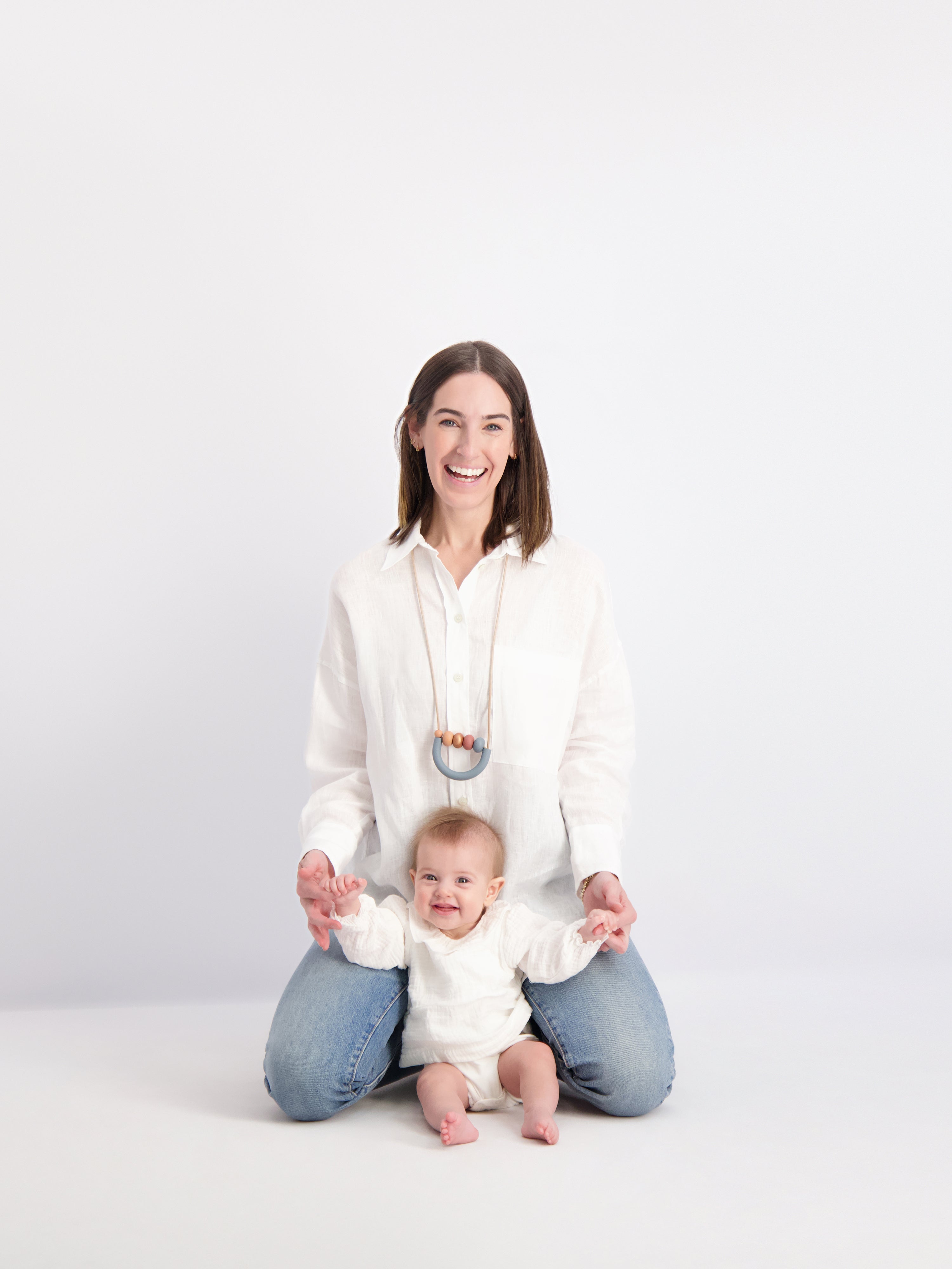 Mom and her baby sitting on the floor and smiling at the camera and holds her baby's hands. Mom is wearing our Loop Teething Necklace in Aegean colorway.